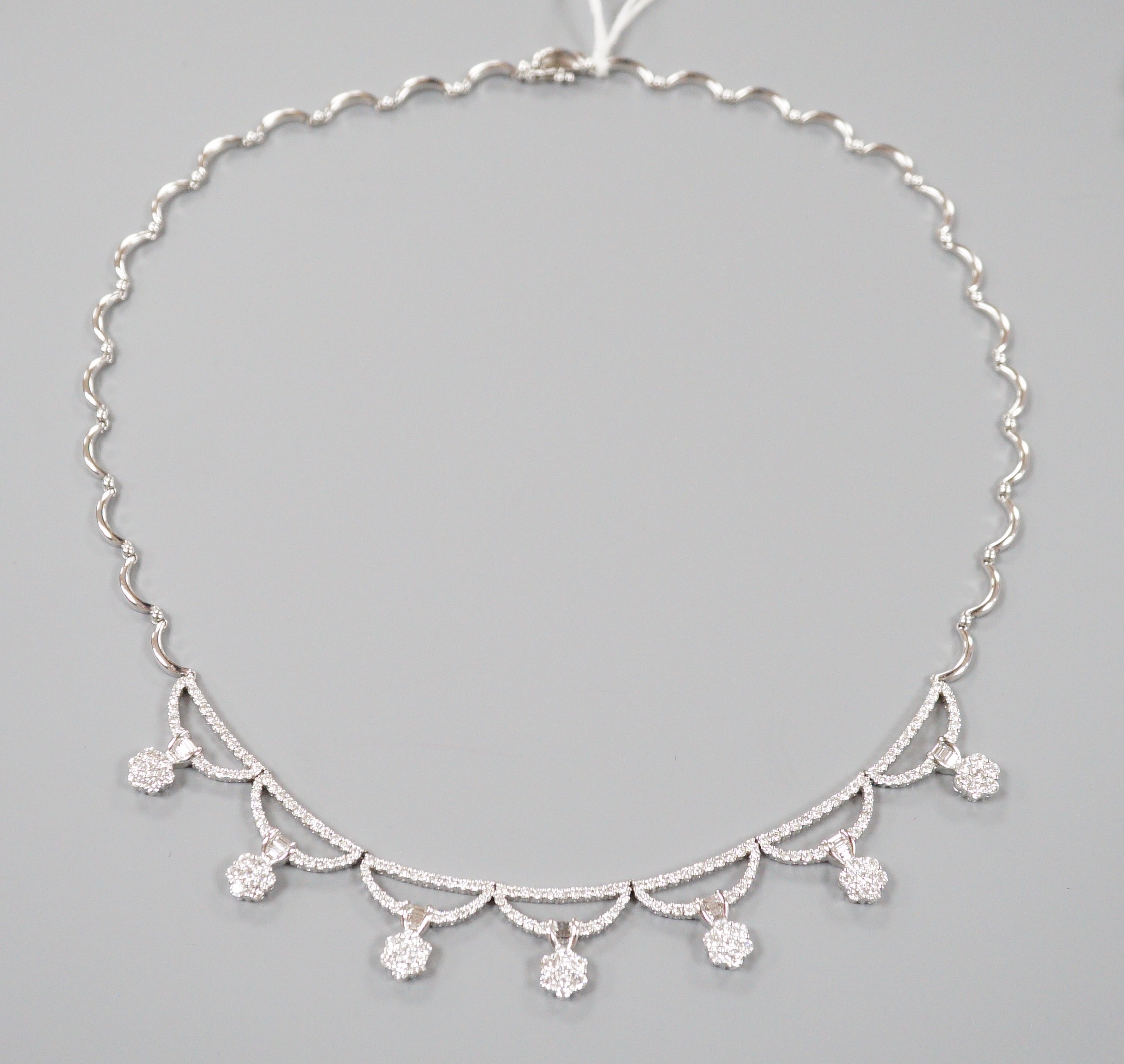 A modern 750 white metal and septuple baguette and round cut diamond cluster set curved link fringe necklace, 43cm, gross weight 26.6 grams